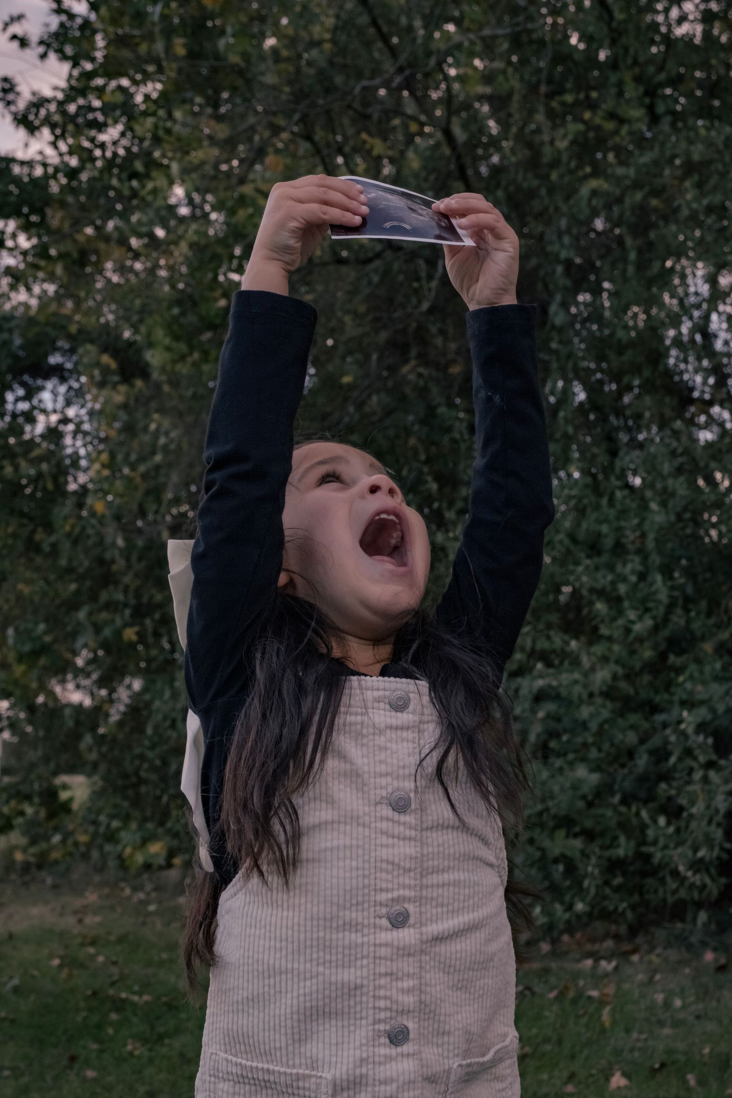 A child playing outdoors reaches up towards the sky while holding something in their hands.