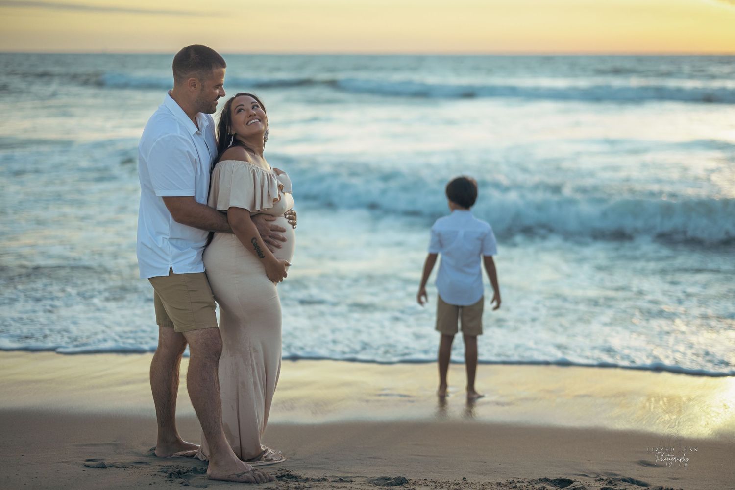 Pregnant couple embraces on a beach at sunset while a child plays near the ocean waves.