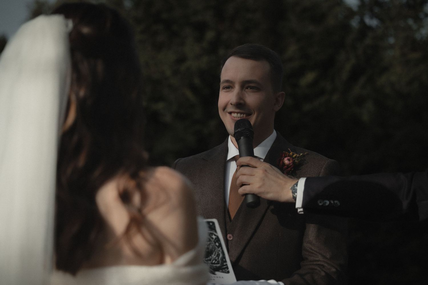 A groom delivers a heartfelt wedding speech at an outdoor ceremony while holding a microphone.
