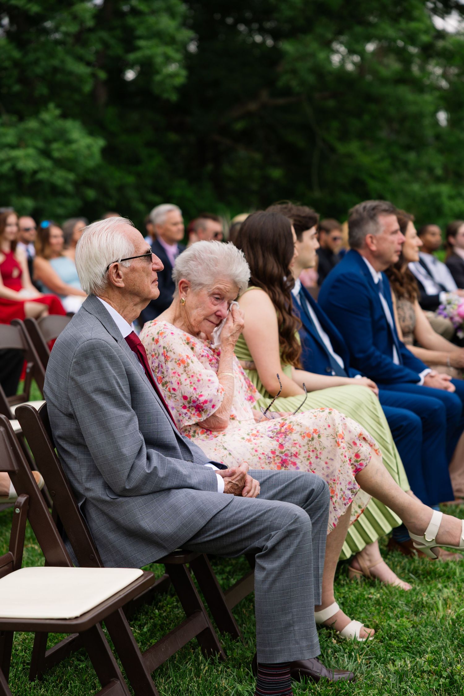 Wedding guests seated on wooden chairs outdoors share a laugh during an outdoor garden ceremony.