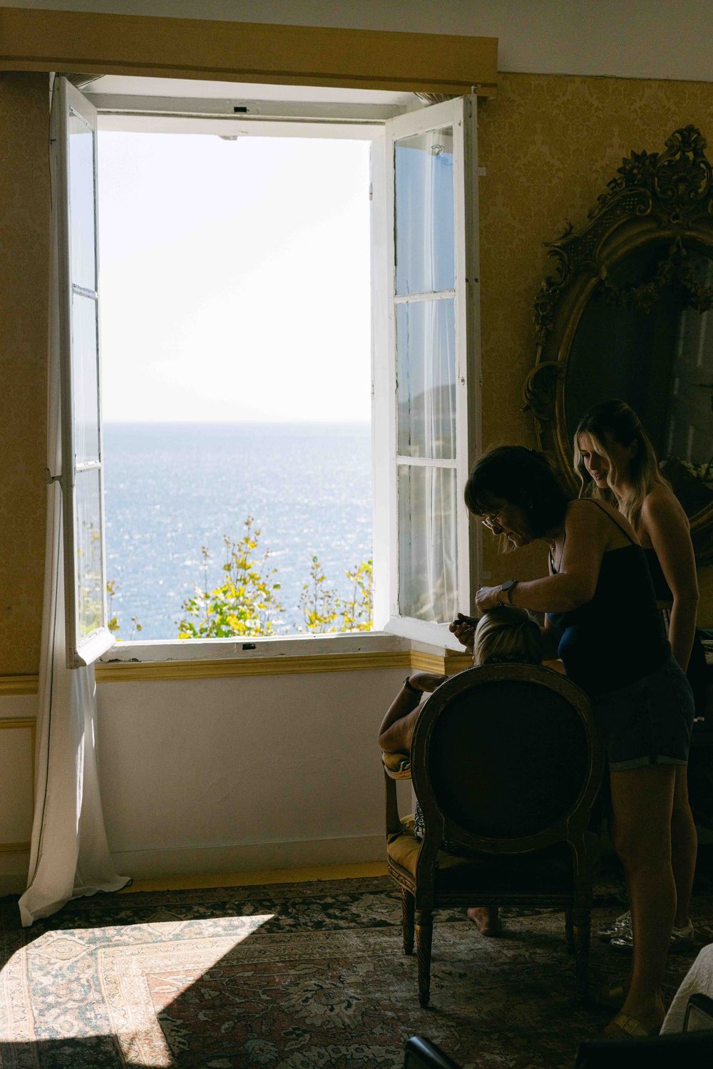 A sunlit room with open windows overlooking the ocean, featuring elegant vintage furniture and ornate decor.