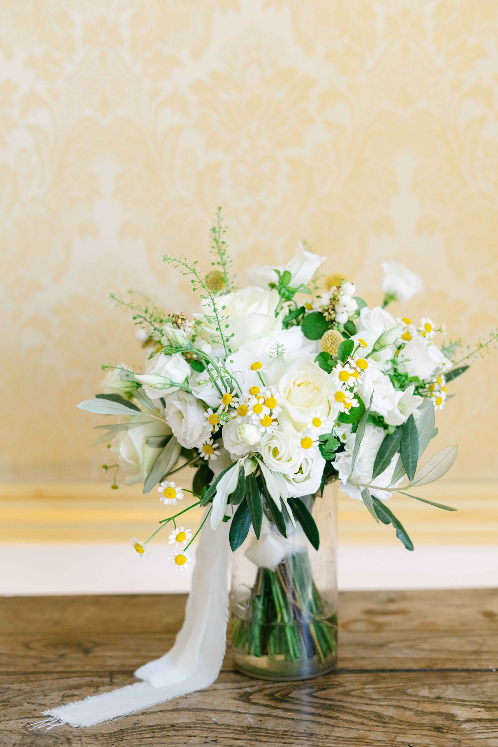 White daisies and greenery arranged in a clear glass vase on a rustic wooden surface against a cream colored wall.