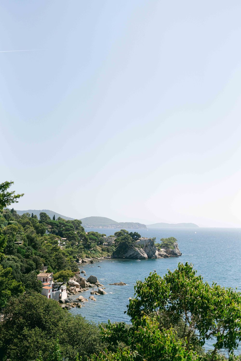 Scenic coastal view of a rocky Mediterranean shoreline with blue waters and lush green vegetation against mountains.