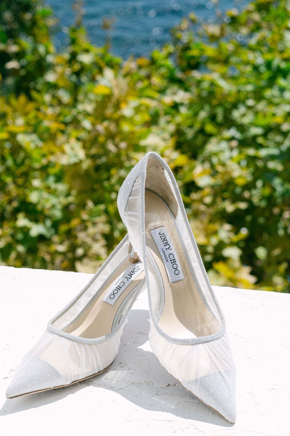 White satin bridal heels displayed against a blurred natural background with water and greenery.