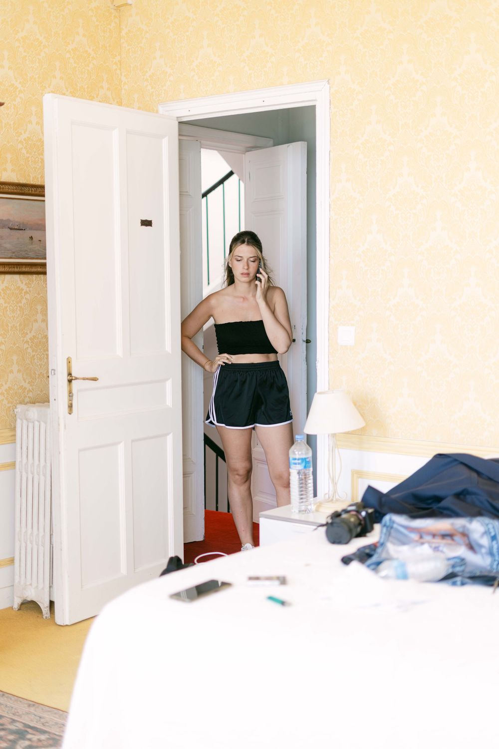 Interior bedroom scene with a white door frame and yellow walls, showing scattered clothing and a lamp on furniture.