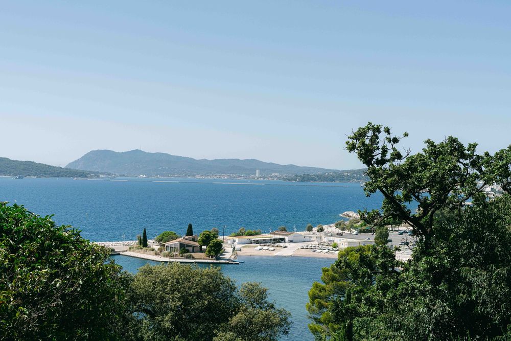 Scenic coastal view of a blue bay surrounded by green trees and distant mountains on a sunny day.