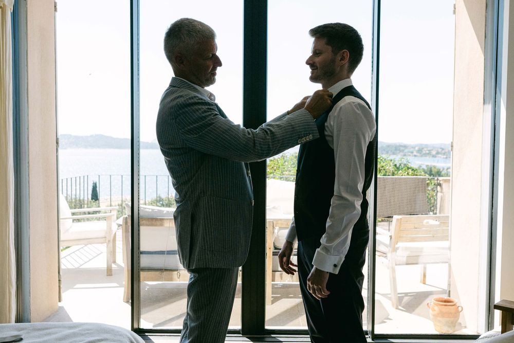 Two people in formal attire stand by large windows in an elegant room with water views in the background.