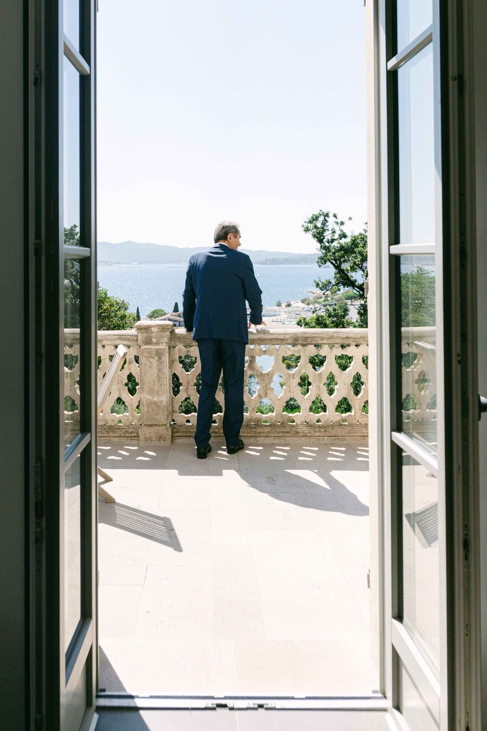 Through open French doors, a person stands on a balcony with ornate stone balustrade overlooking a scenic mountain view.