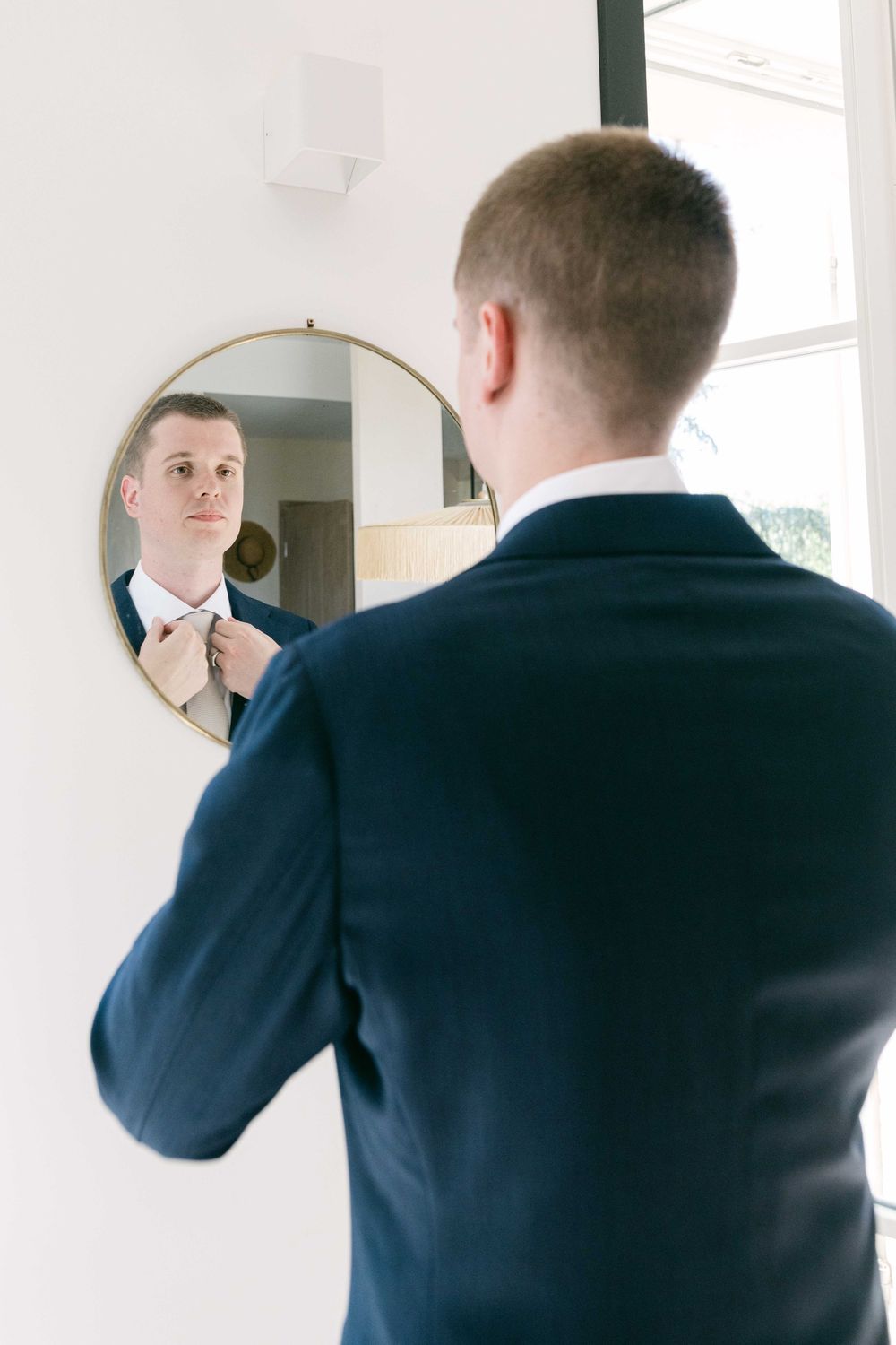 Man in a navy suit jacket looks at his reflection in a mirrored wall while adjusting his appearance.