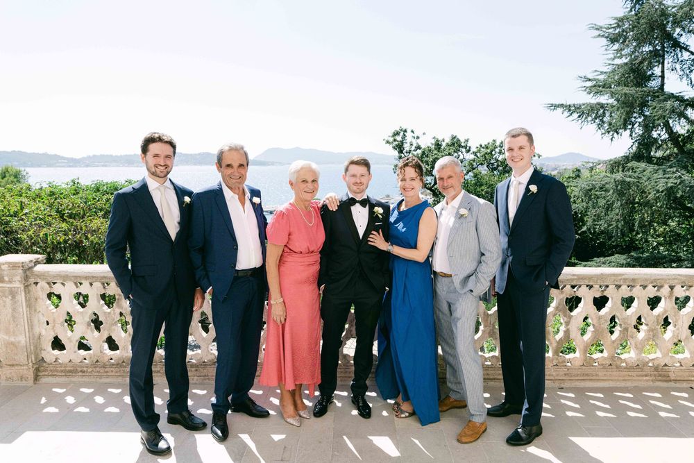 Wedding party members pose together on an elegant stone balcony with scenic views of trees and gardens in the background.