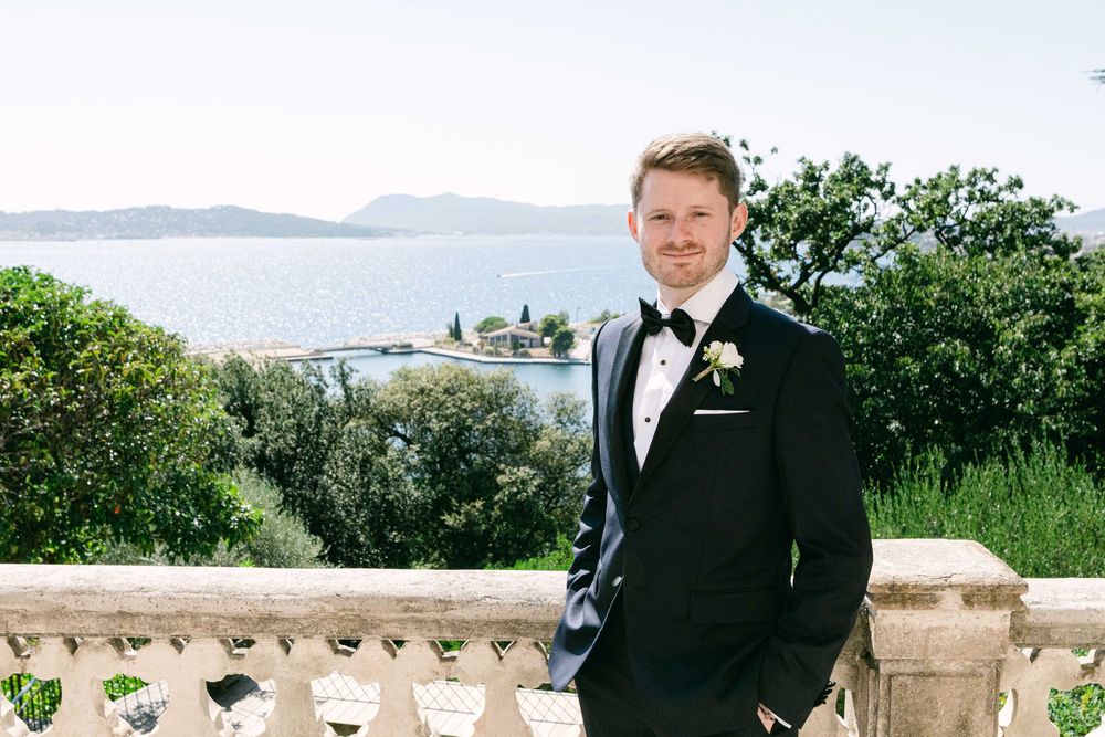A groom in a classic black tuxedo and bowtie stands on a stone balcony overlooking a scenic coastal view.
