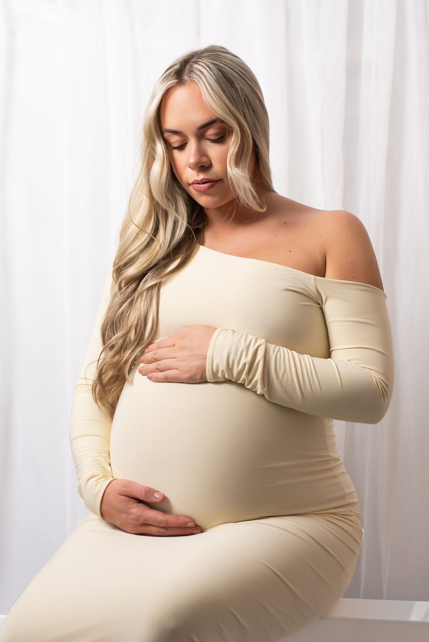 Woman in beige off-shoulder dress poses against white curtain background in maternity photoshoot series.