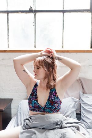Person in floral print top adjusts their hair while sitting on bedding near a window.