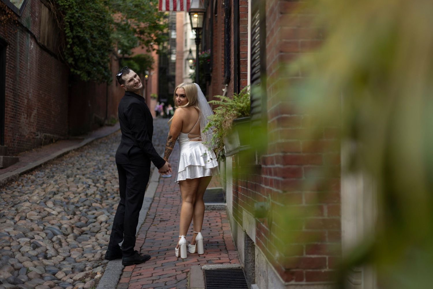 A couple stands together on a historic cobblestone street with brick buildings and ivy in Boston's Beacon Hill.