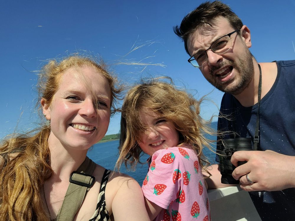 A family takes a sunny outdoor selfie together against a bright blue sky on a windy day.