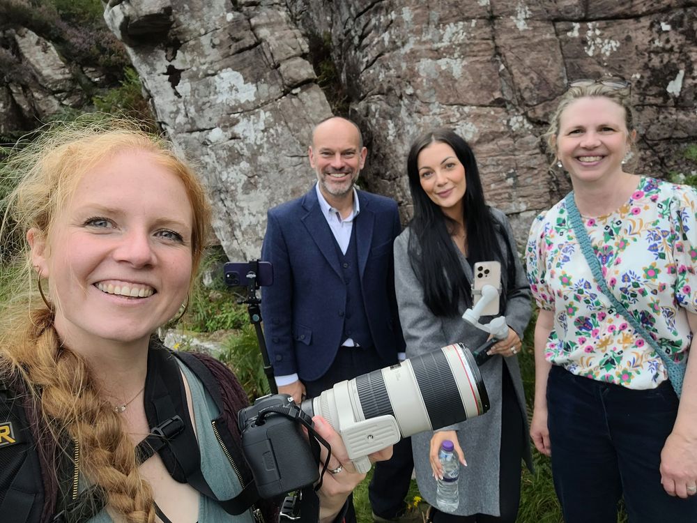 A photographer with professional camera equipment poses with three others in front of a rocky cliff face outdoors.
