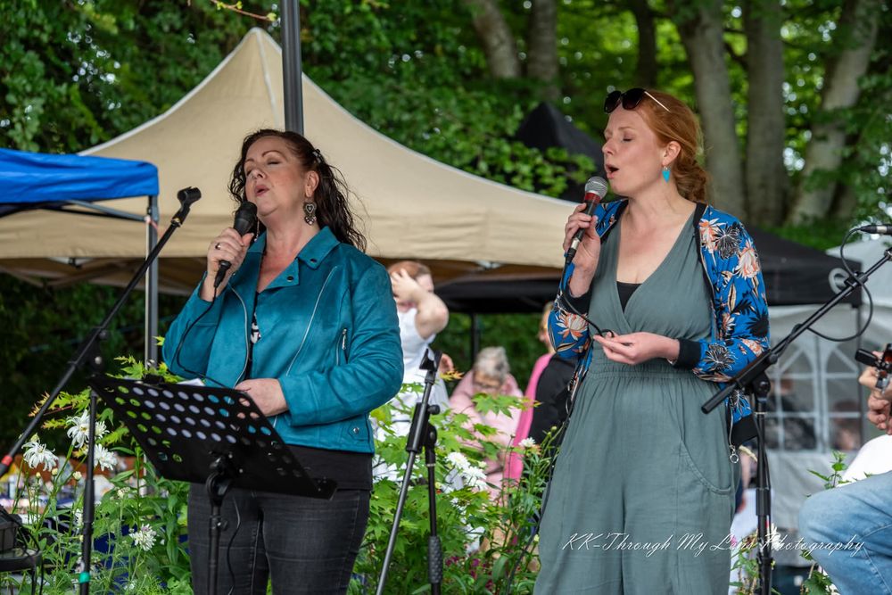 Two singers perform together outdoors under a canopy tent with microphones and music stands in a wooded setting.
