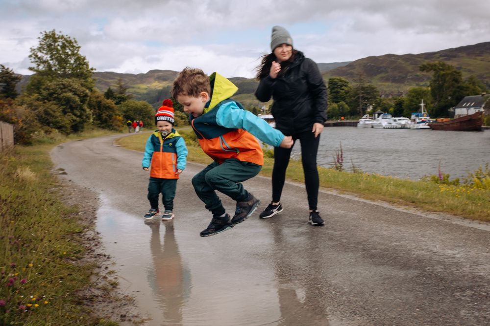 People splash and jump through puddles on a wet path near a scenic mountain landscape.