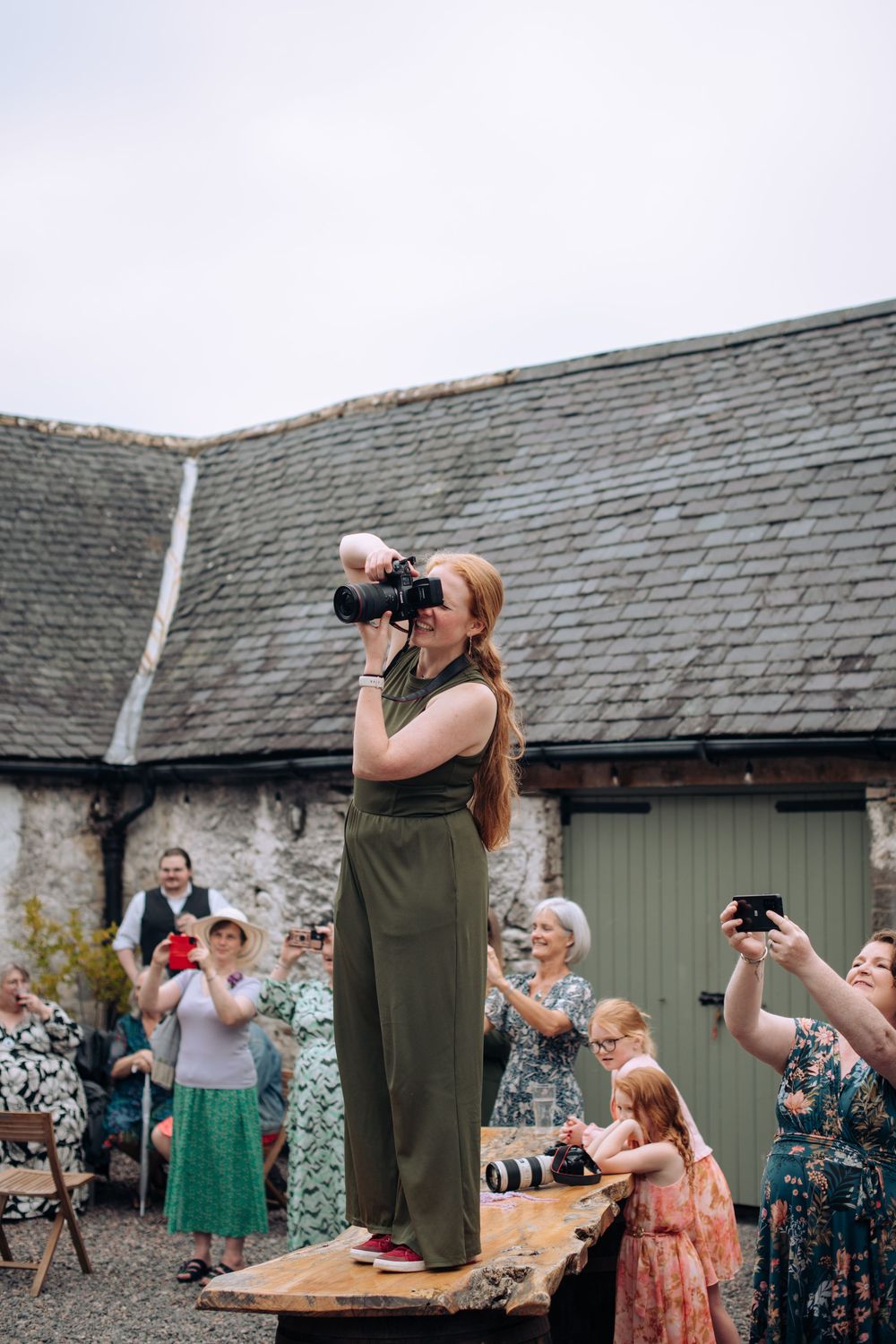 Wedding guests gather outdoors for drinks and celebration at a rustic venue with slate roof buildings.