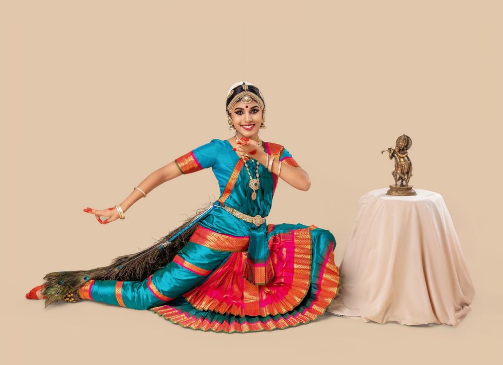 Classical Indian dancer in vibrant turquoise and pink costume performs graceful poses on beige backdrop.