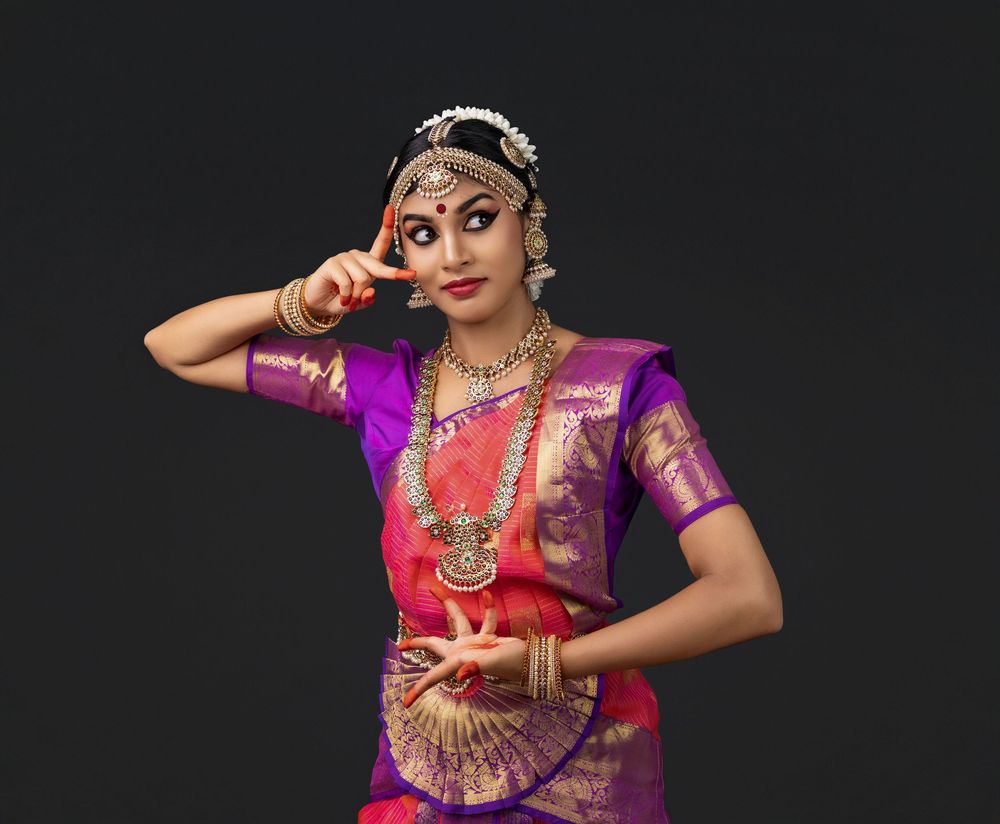 Indian classical dancer performs traditional dance wearing vibrant purple and pink sari with ornate jewelry against dark background.