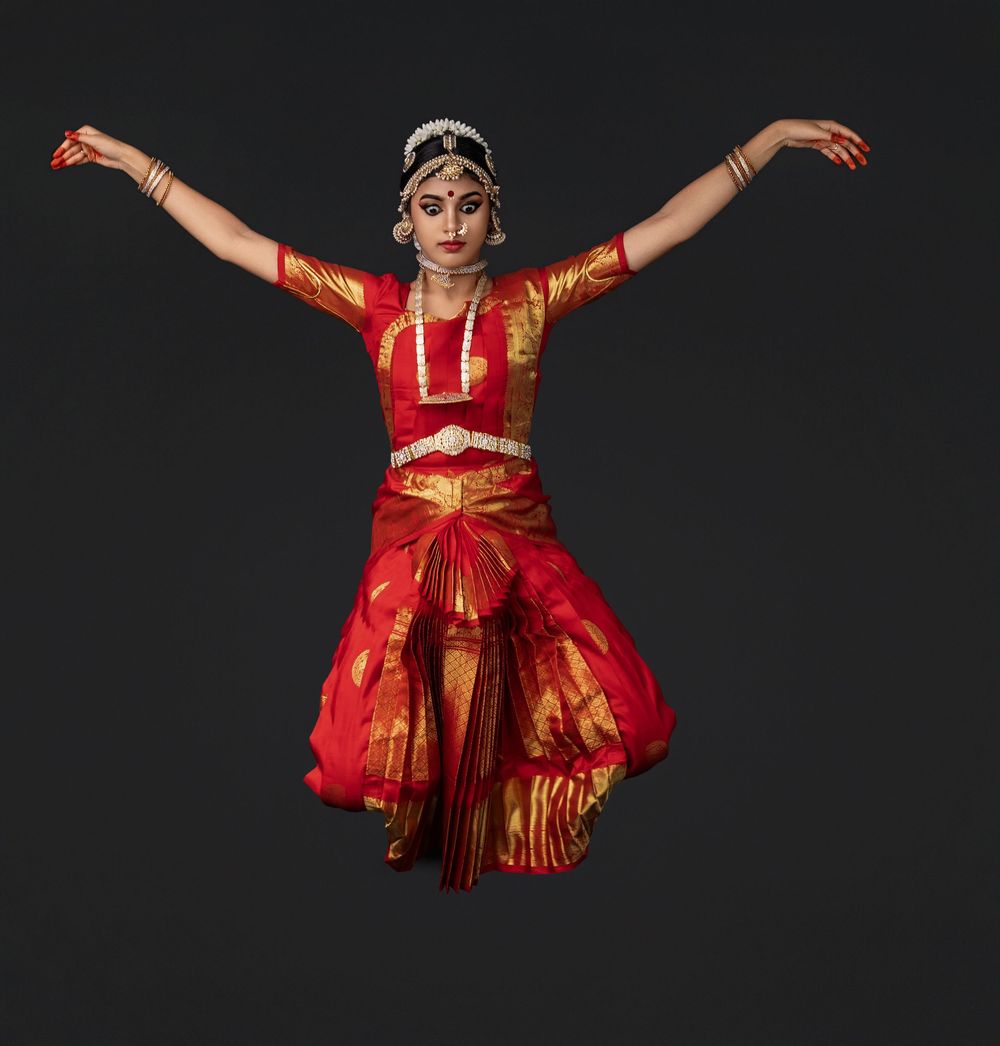 Dancer in vibrant red and gold traditional Indian dance costume performs graceful movements with outstretched arms.