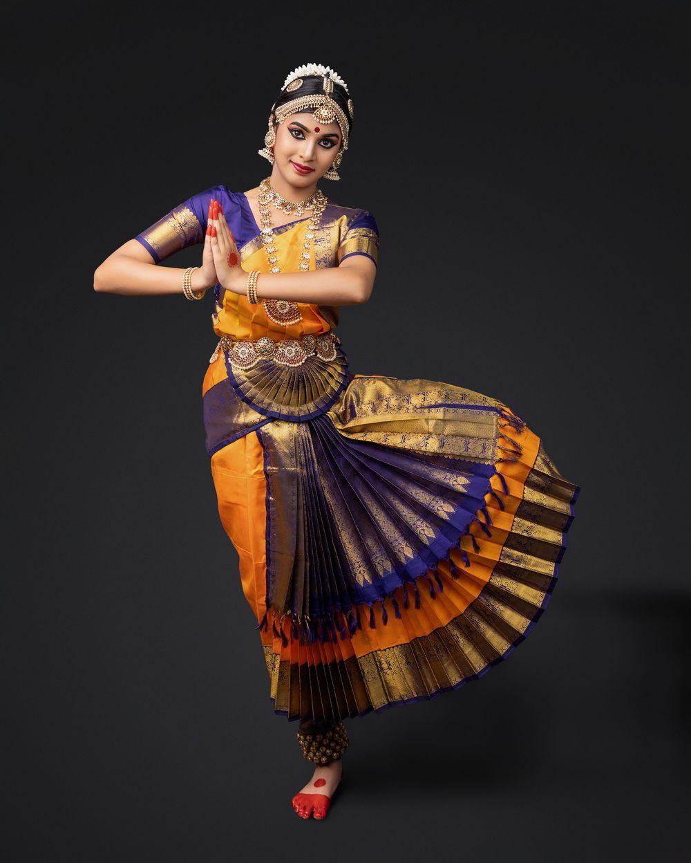 Dancer performs traditional Indian classical dance Bharatanatyam in vibrant orange and purple costume against dark backdrop.