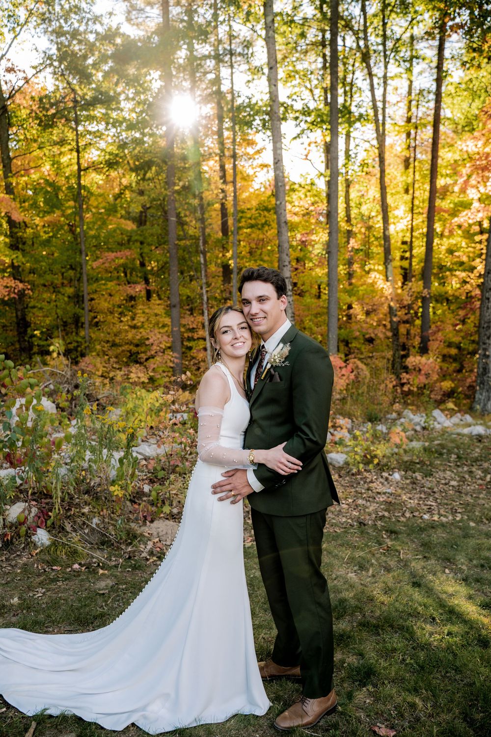 A wedding couple embraces in front of autumn trees with golden sunlight streaming through the forest.
