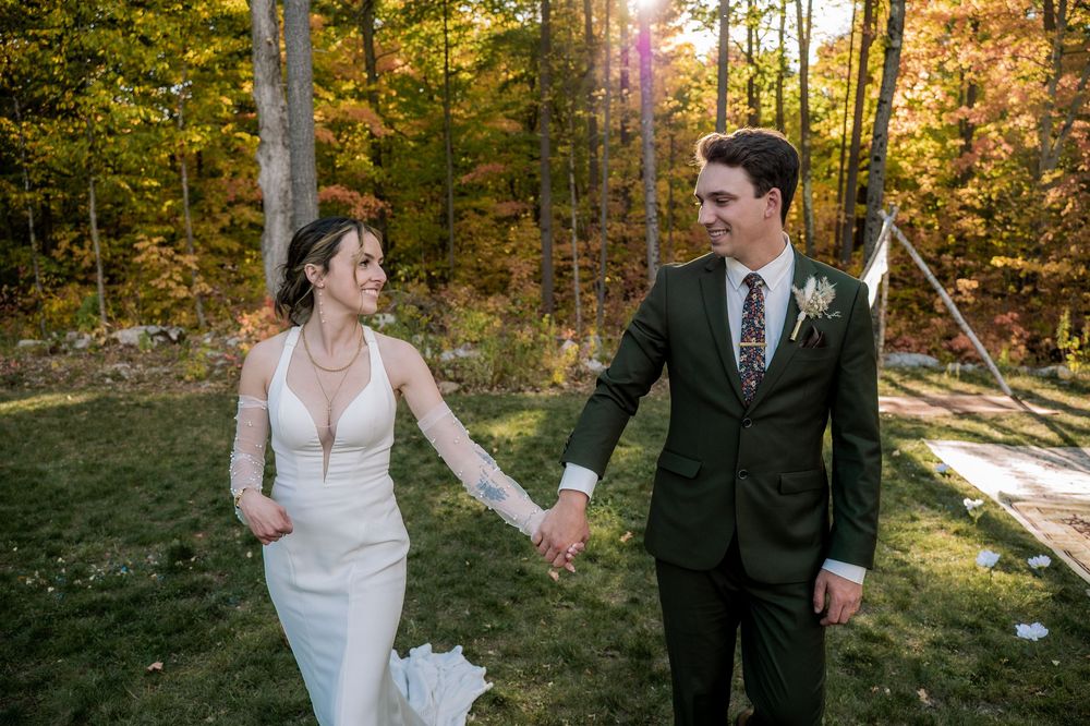A couple holding hands and walking through a sunlit autumn forest during an outdoor wedding ceremony.