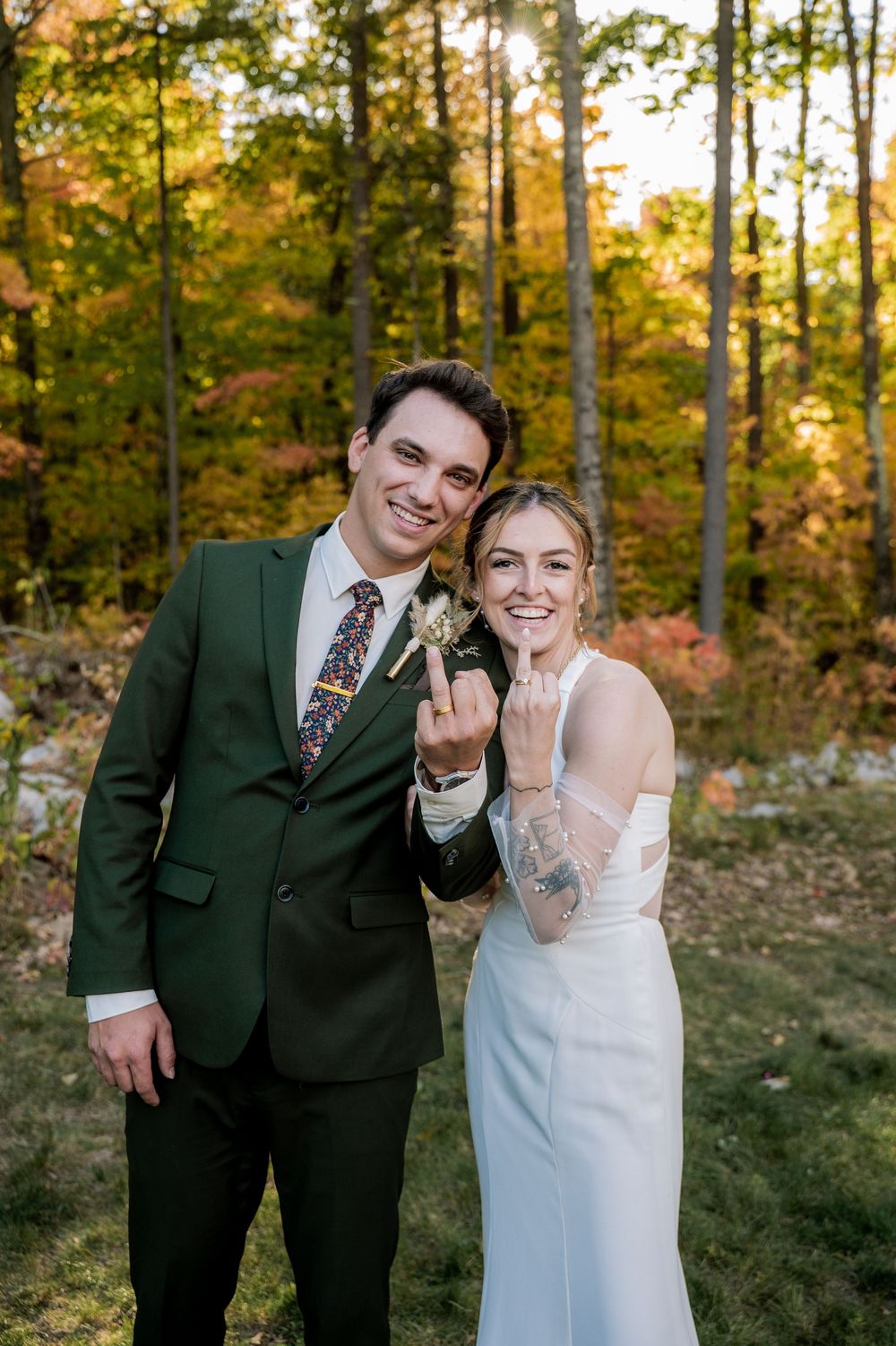 A couple in wedding attire stands in a forest during golden hour showing off their wedding rings.