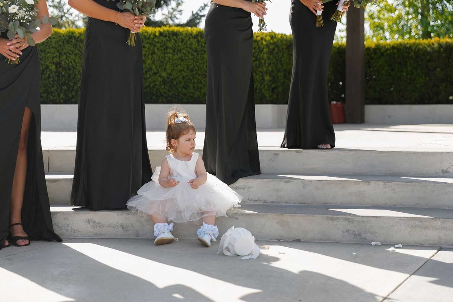 Adorable flower girl in white tutu dress sitting on steps beside bridesmaids in black gowns.