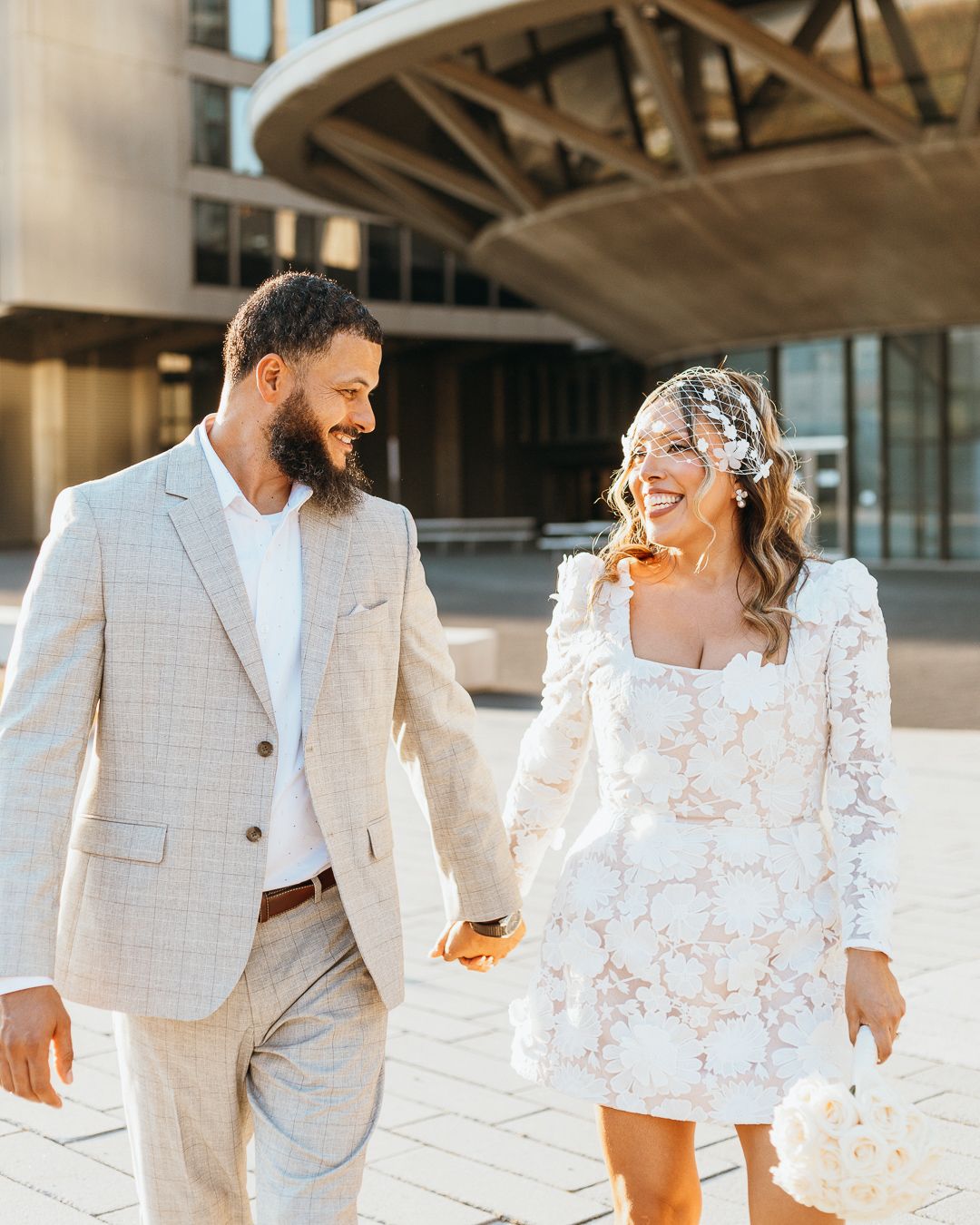 Couple in elegant white outfits walking and laughing together in an urban setting.