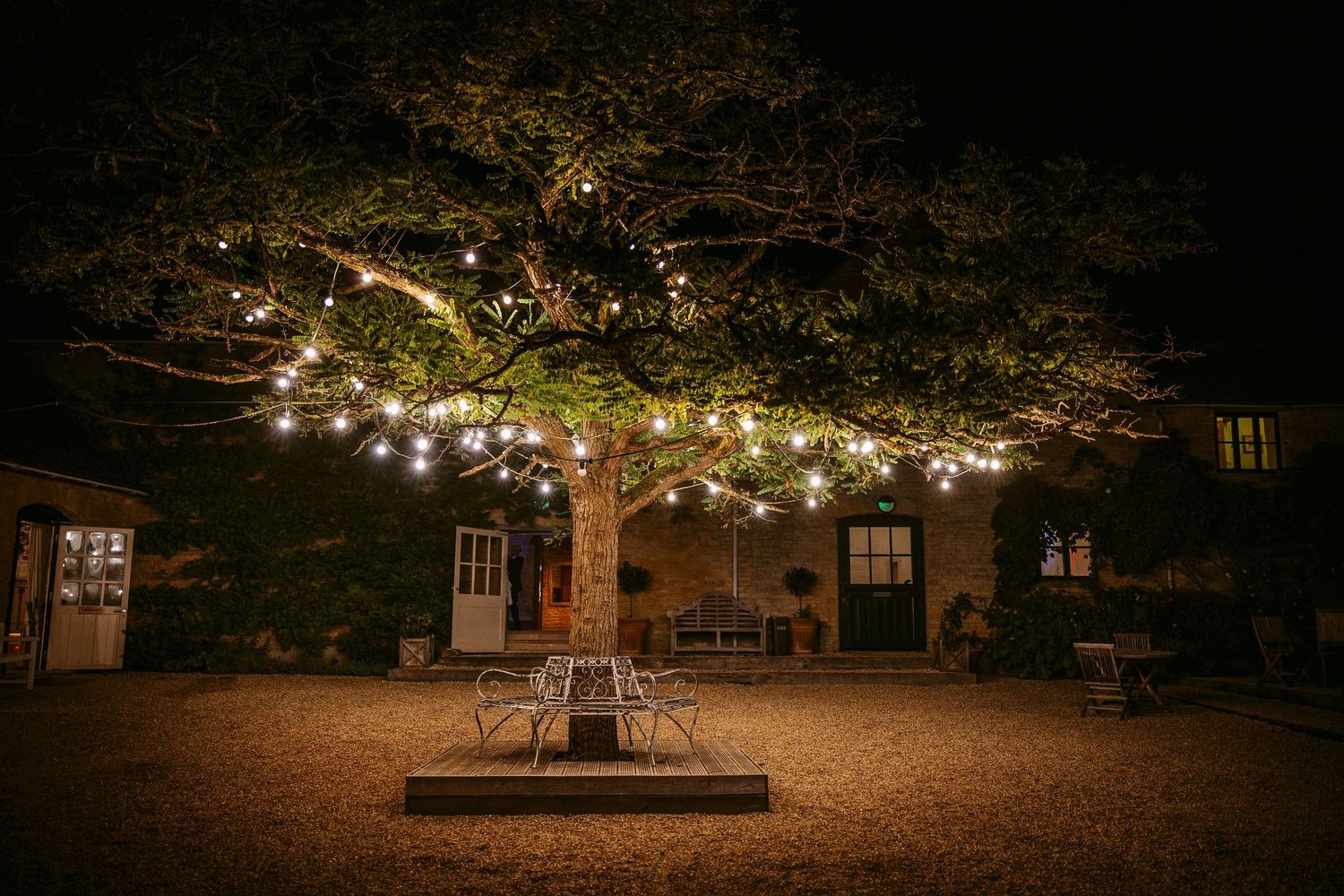 String lights illuminate a rustic outdoor seating area with wooden benches and tables at night.