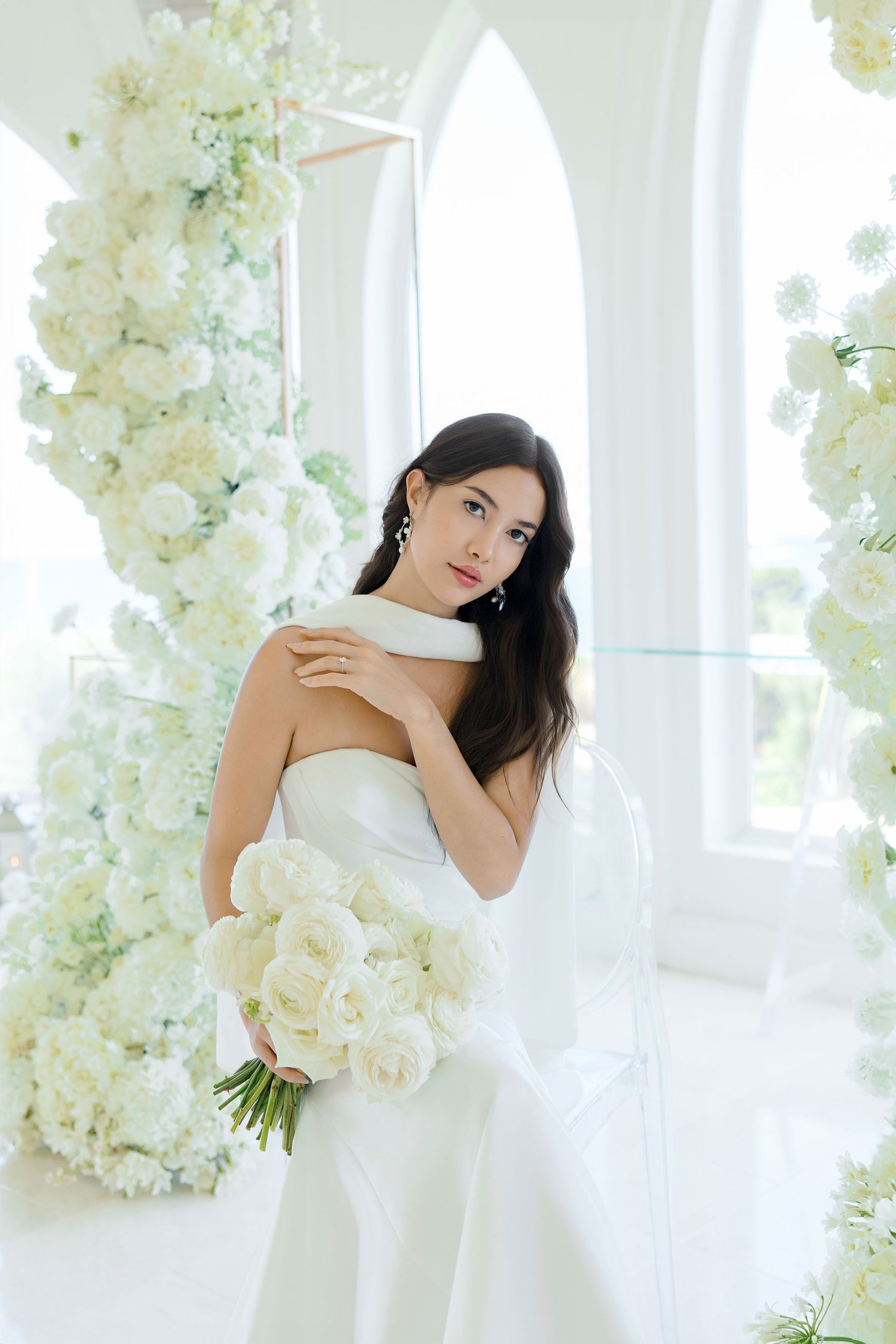 A bride in a white strapless gown poses elegantly among cascading white floral arrangements in a bright studio setting.