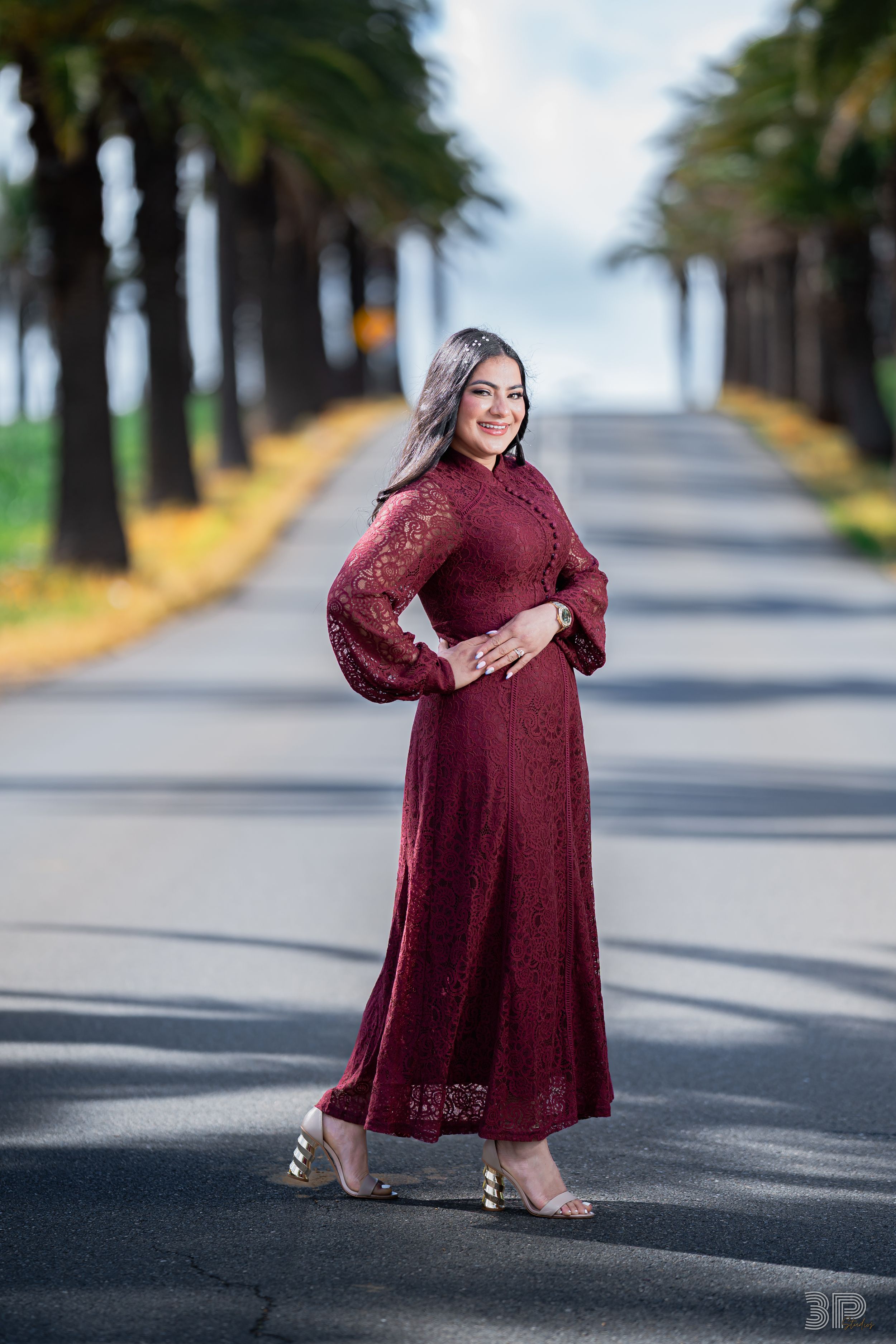 A person in a burgundy lace dress poses gracefully on a tree-lined street with dappled sunlight.