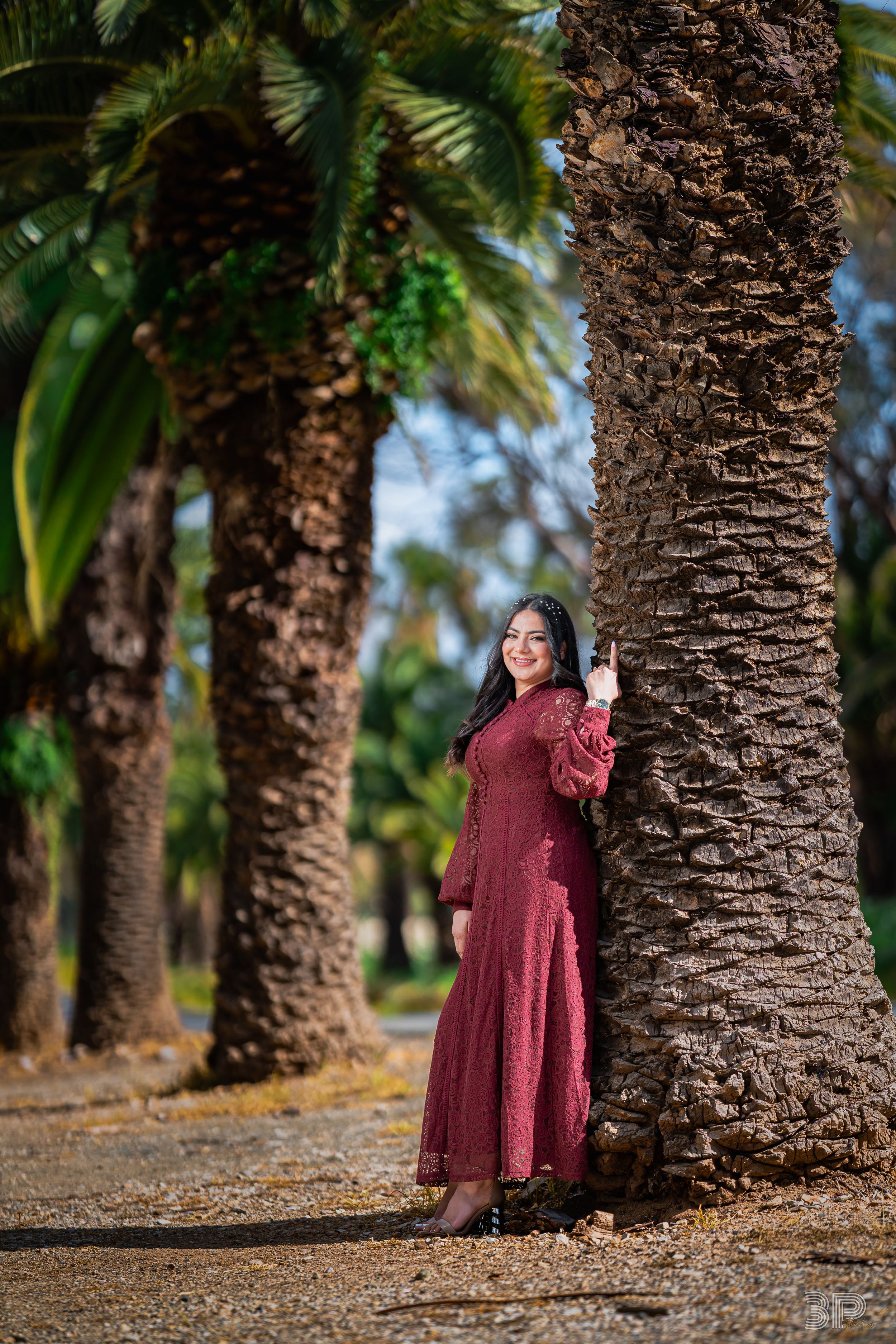 A person in a burgundy dress poses playfully among a row of tall palm trees in an outdoor setting.