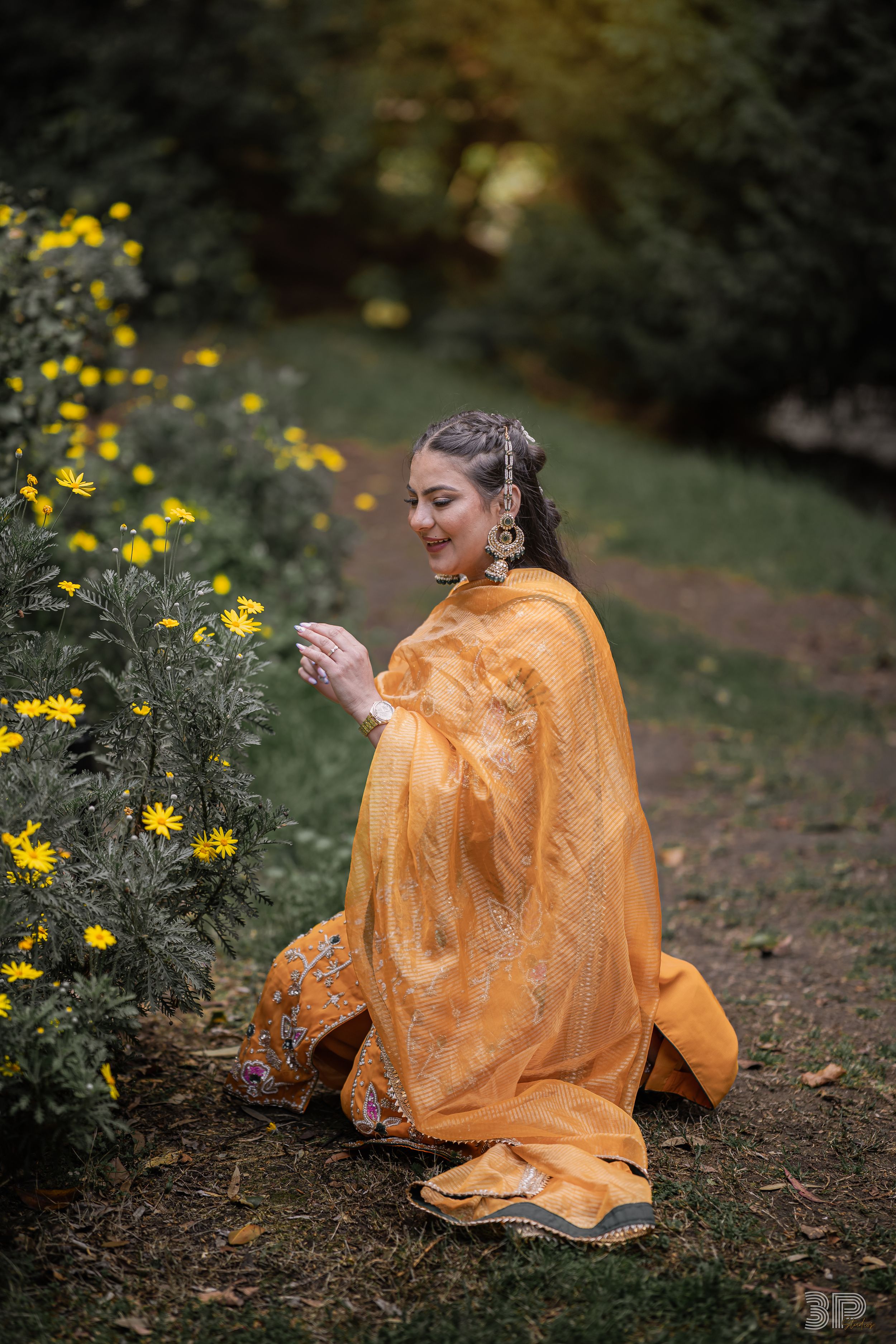 A person in a flowing orange dress sits gracefully among yellow wildflowers in a garden setting.