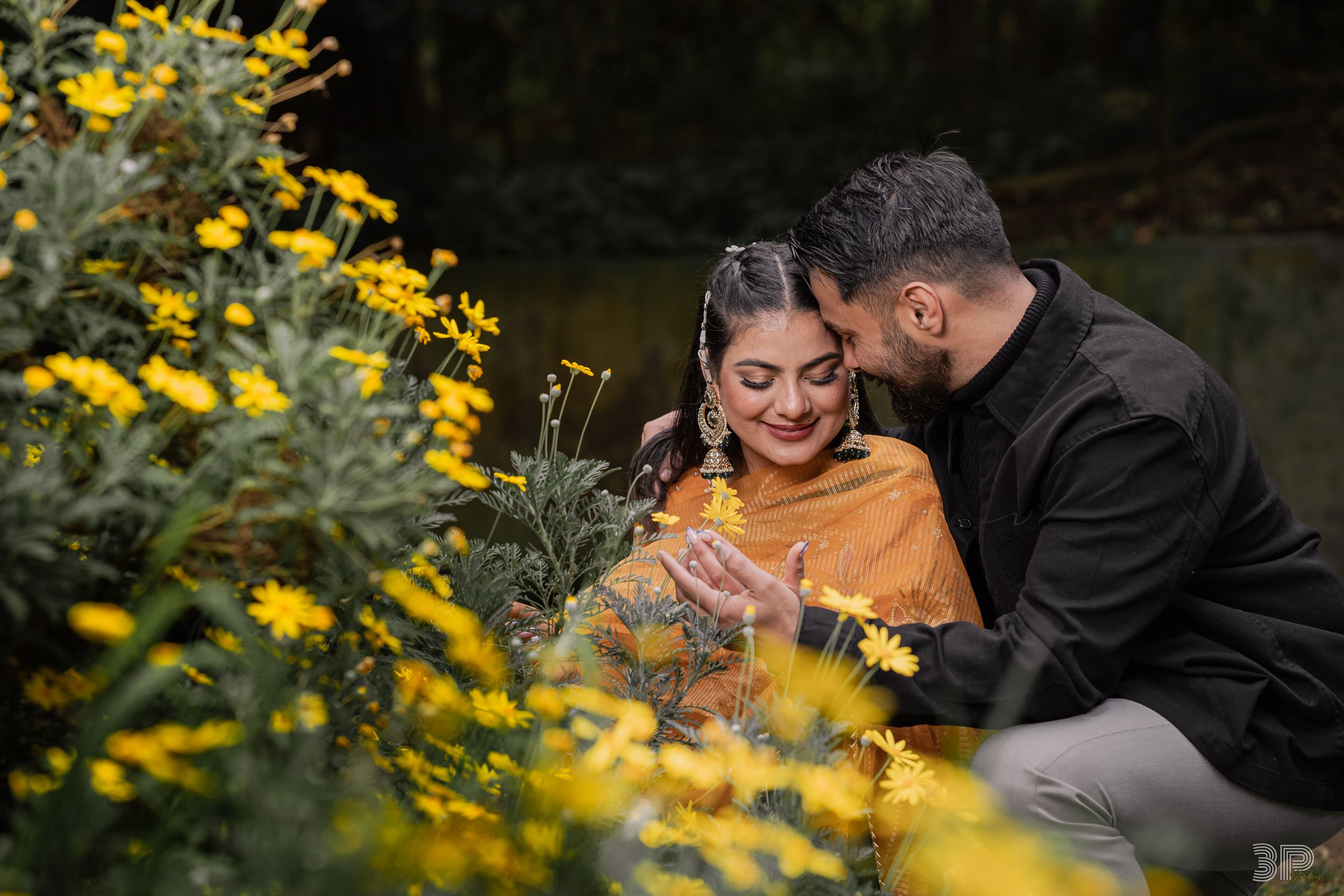 A couple shares an intimate moment surrounded by blooming yellow wildflowers near a tranquil pond.