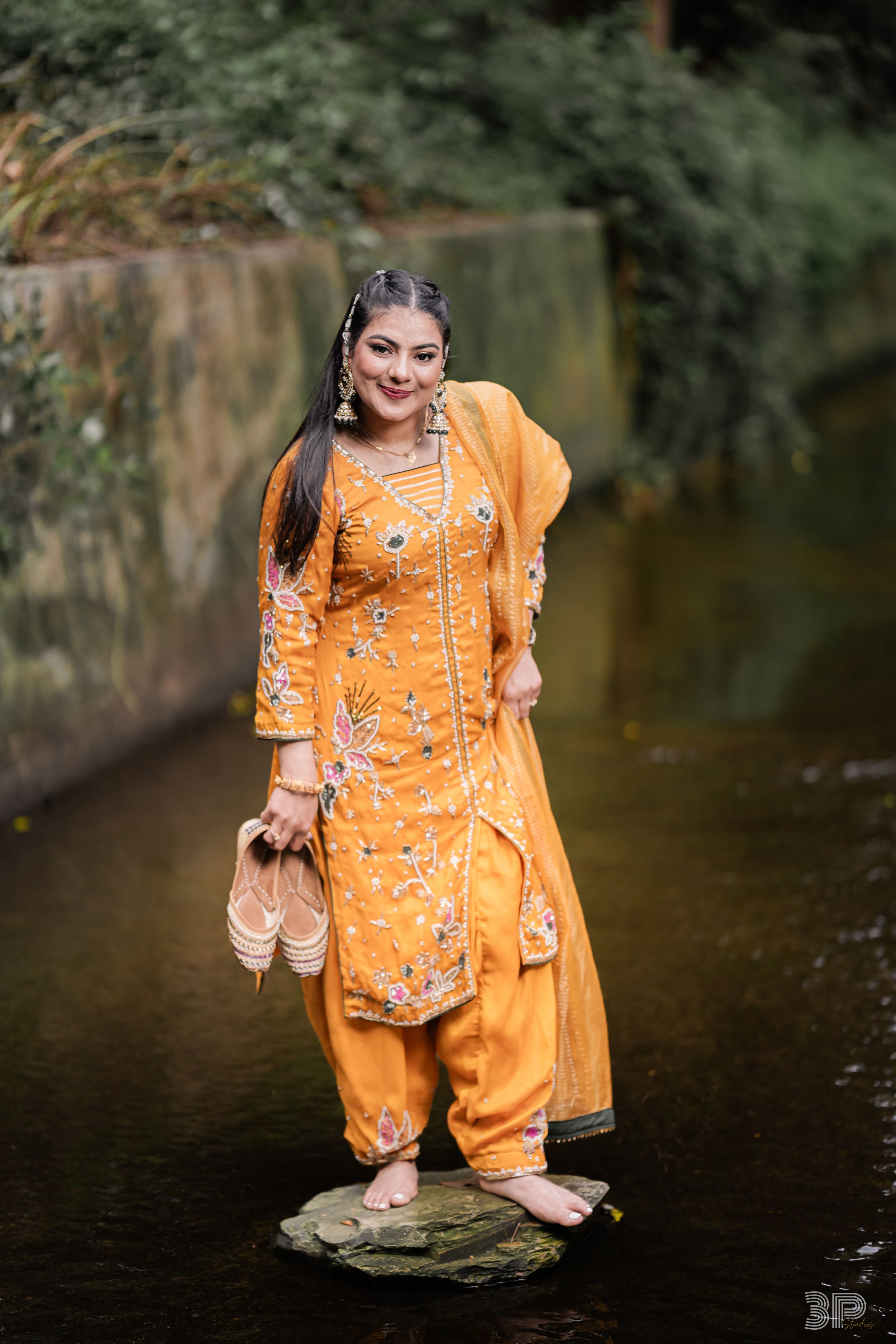 A person in a bright orange traditional Indian dress poses near a reflective pond in an outdoor setting.