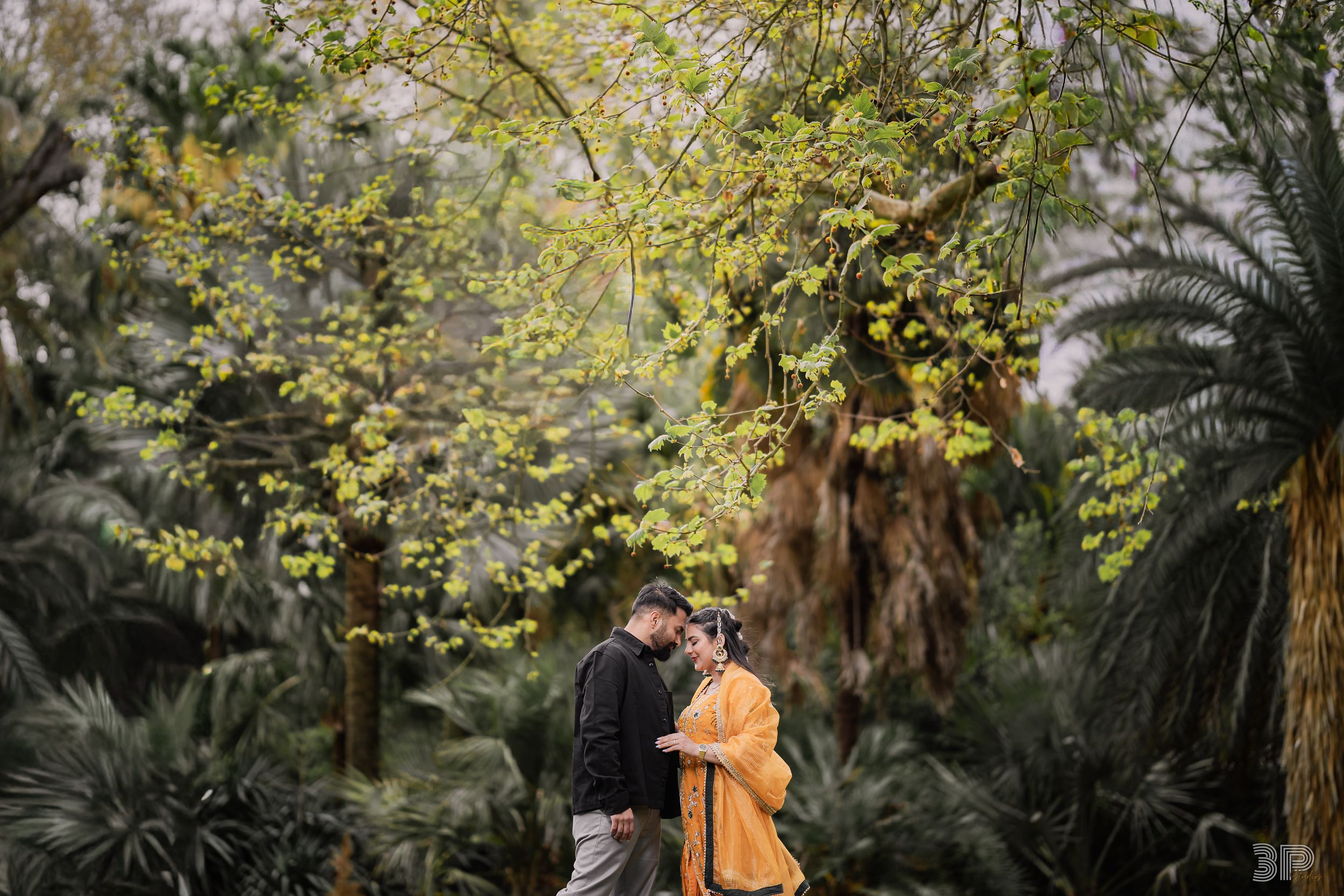 A couple in orange beanies shares a tender moment while walking through a lush autumn garden path.