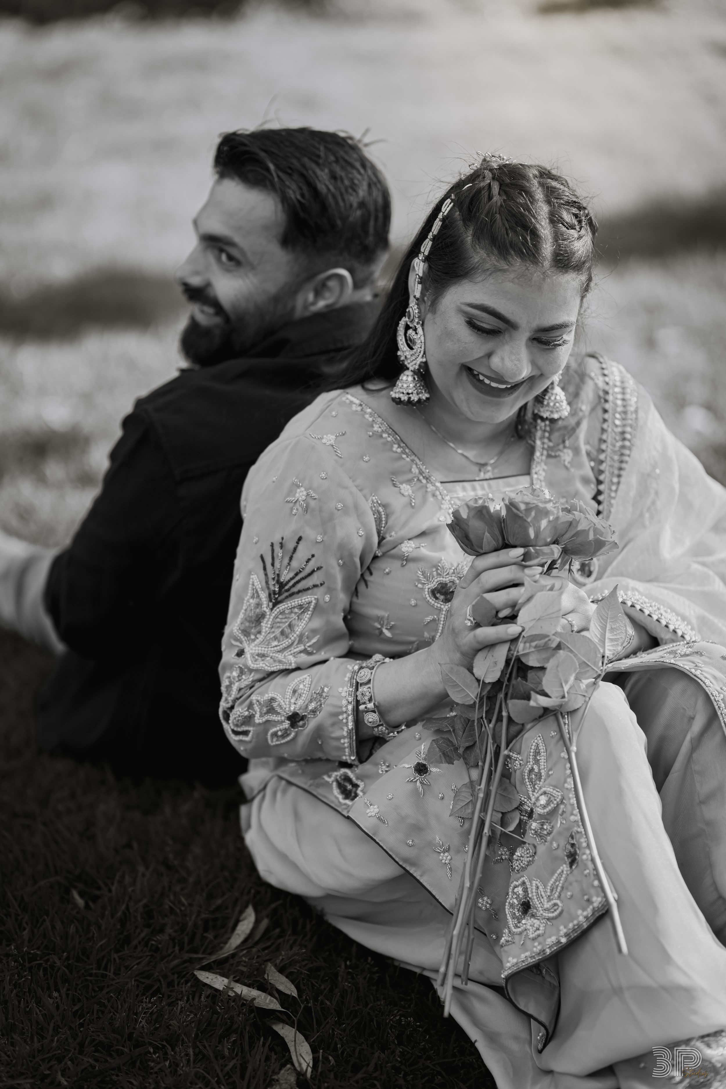 Black and white photo of a couple in traditional South Asian wedding attire sitting together outdoors on grass.