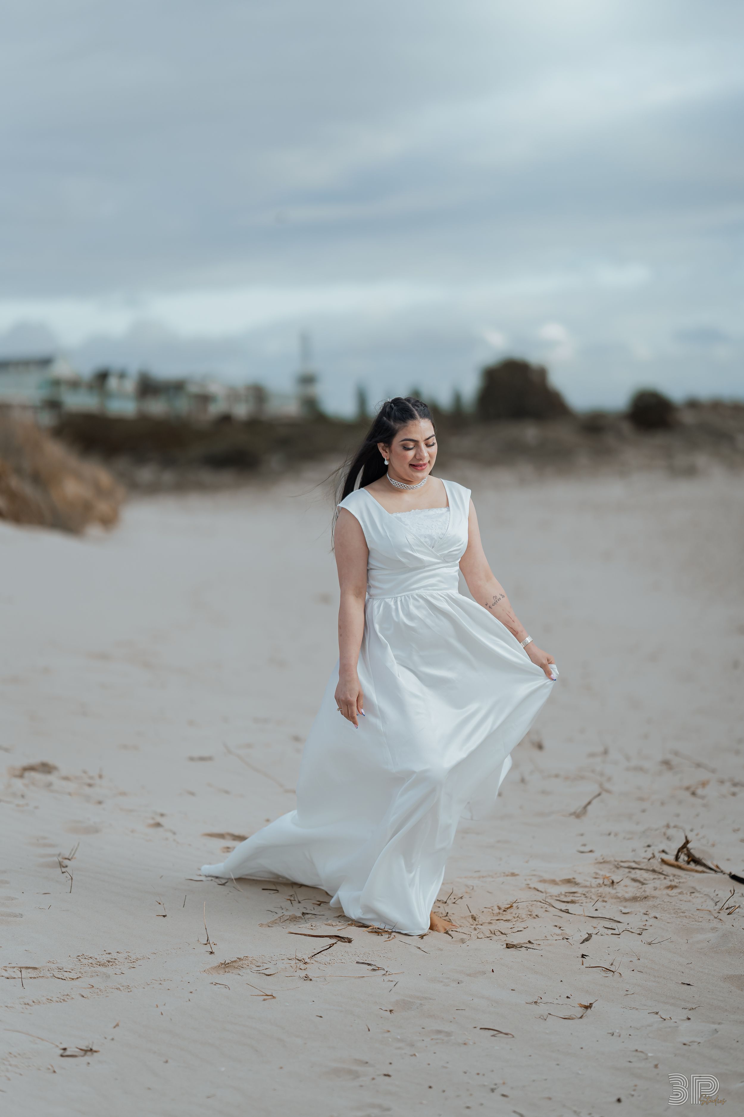 A flowing white dress catches the breeze as someone stands on a sandy beach under cloudy skies at dusk.