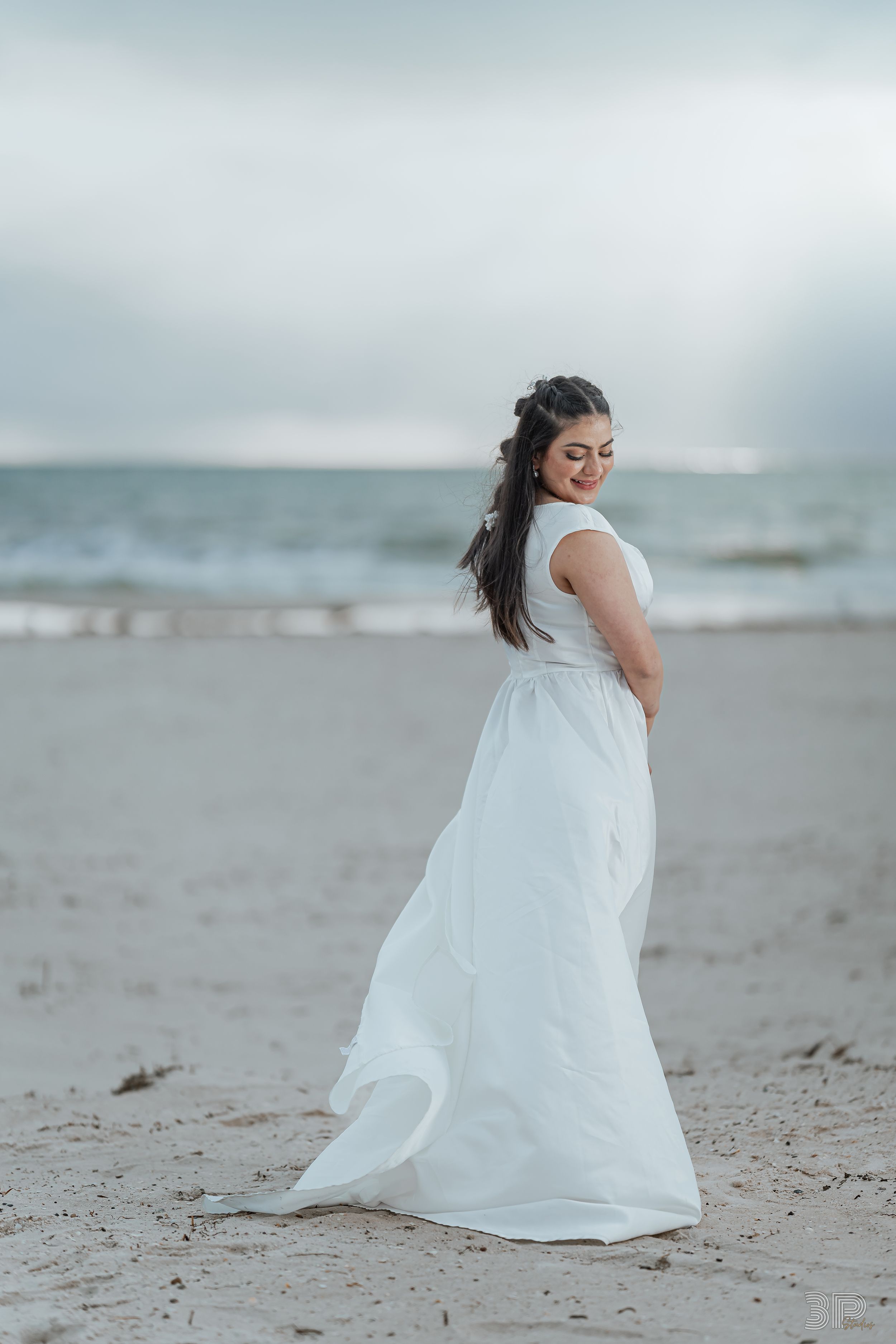 A person in a flowing white dress sits gracefully on a misty beach shoreline in a dreamy coastal setting.