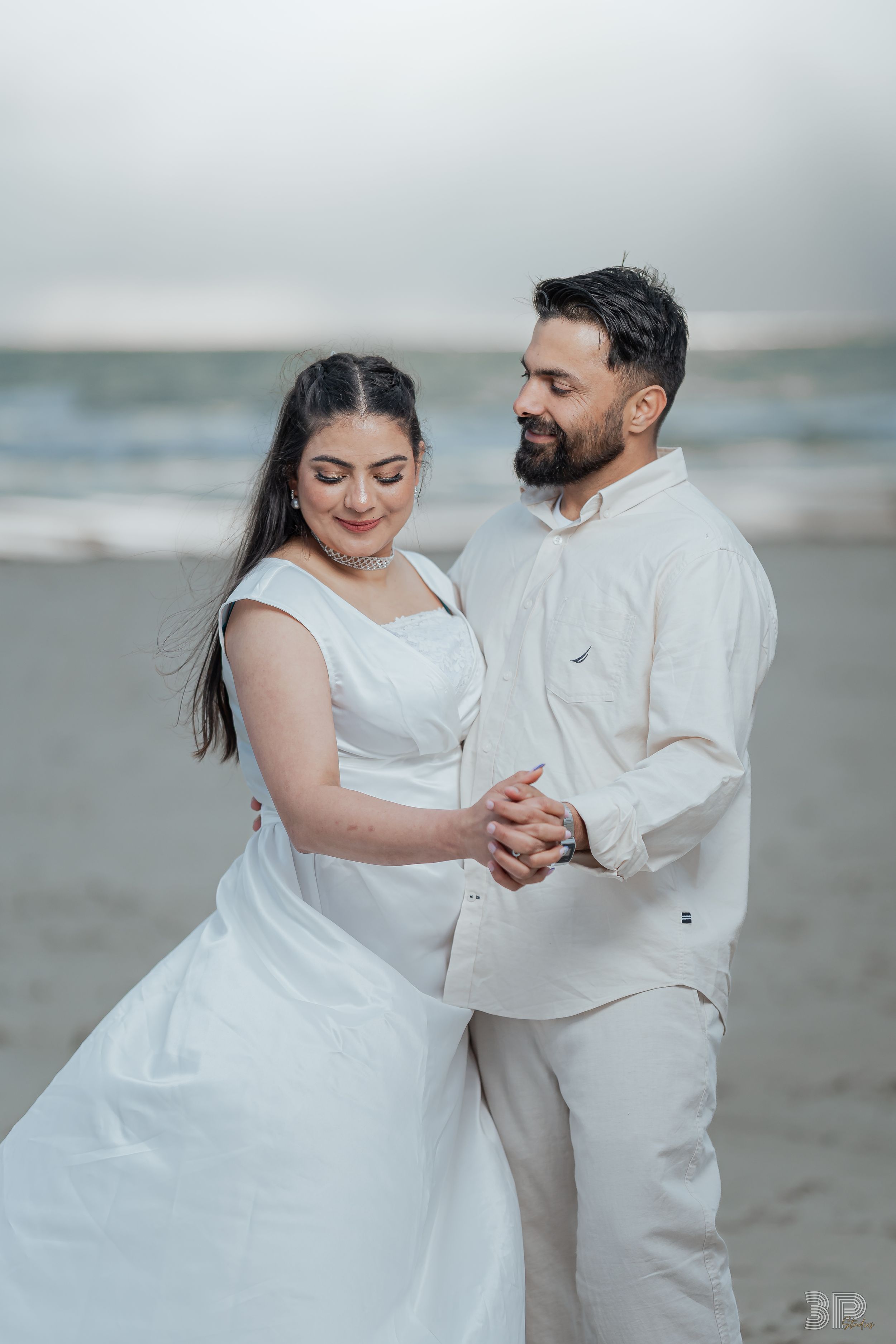 A couple in white outfits dances romantically together on a misty beach during a photoshoot.