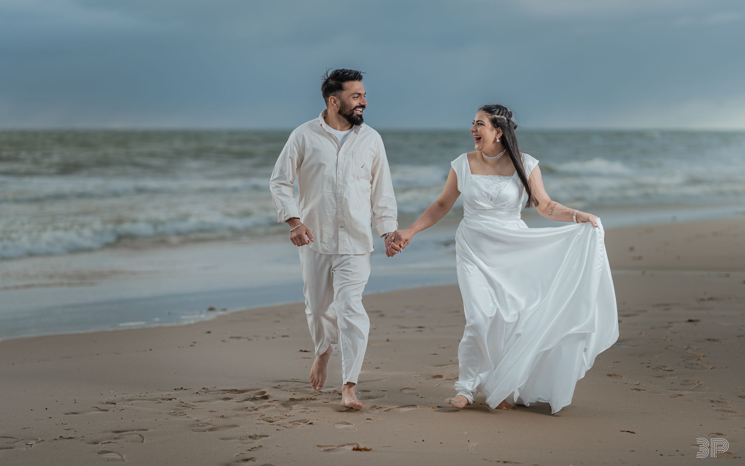 A couple in white attire walks hand in hand along a beach at dusk with waves crashing in the background.