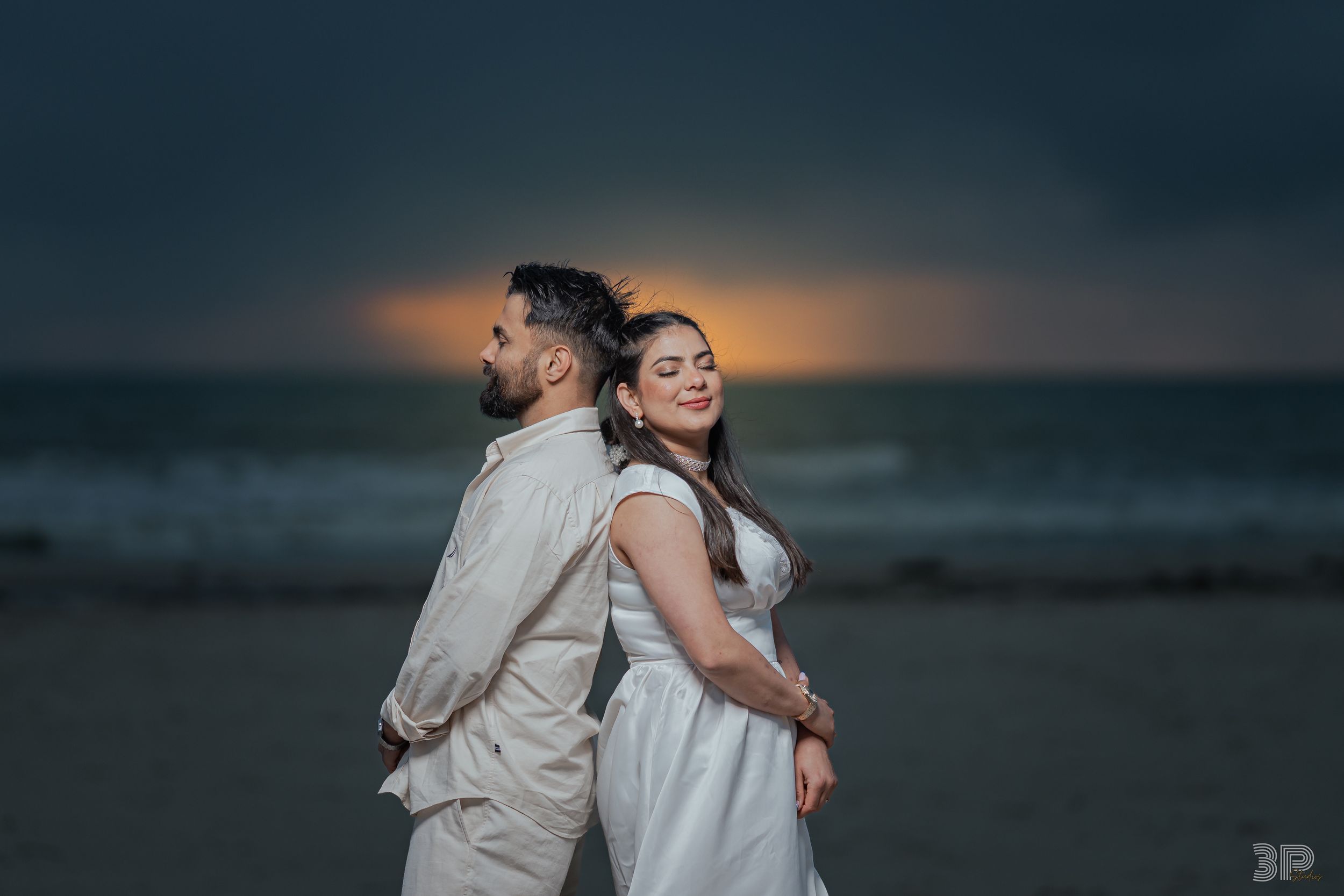 A couple stands back-to-back on a beach at sunset with dramatic moody sky and ocean waves in the background.