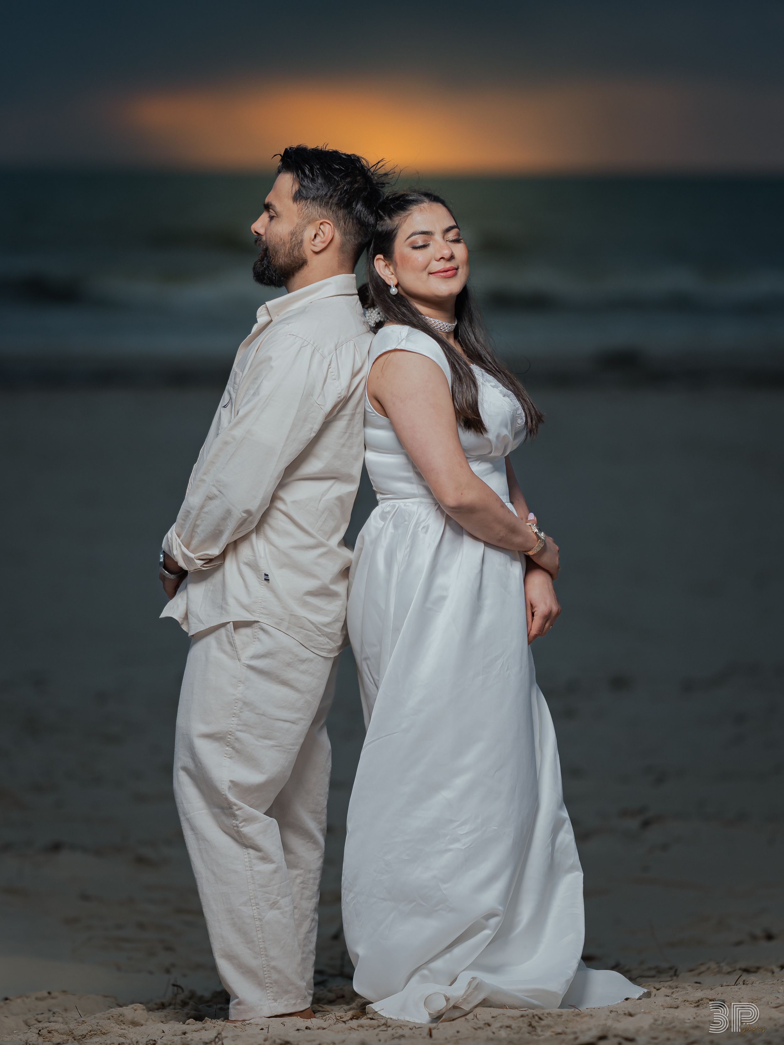 A couple in white outfits stands back-to-back on a beach at sunset creating a romantic silhouette against the ocean.