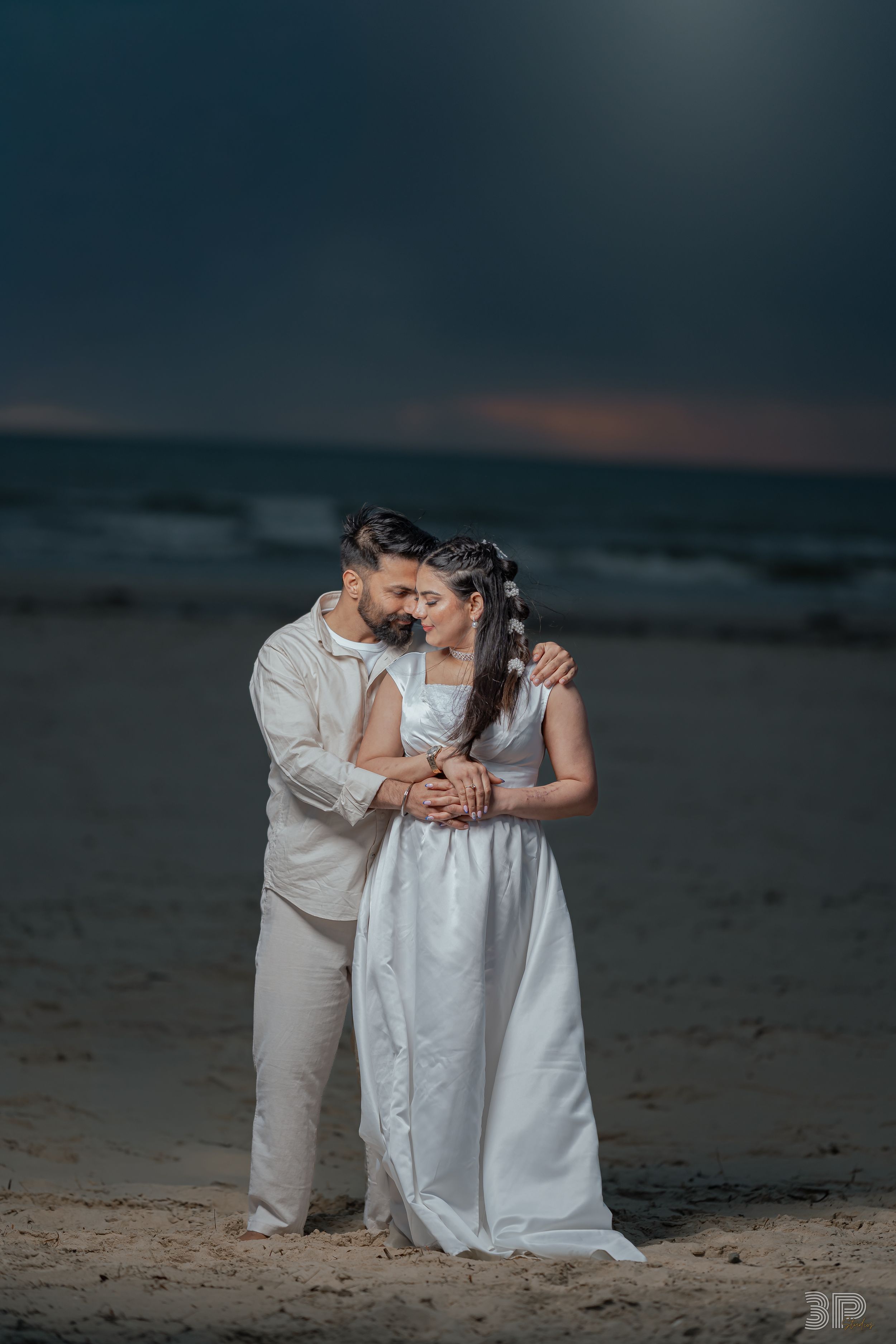Romantic couple embraces on a moody beach at dusk wearing white and beige formal attire.