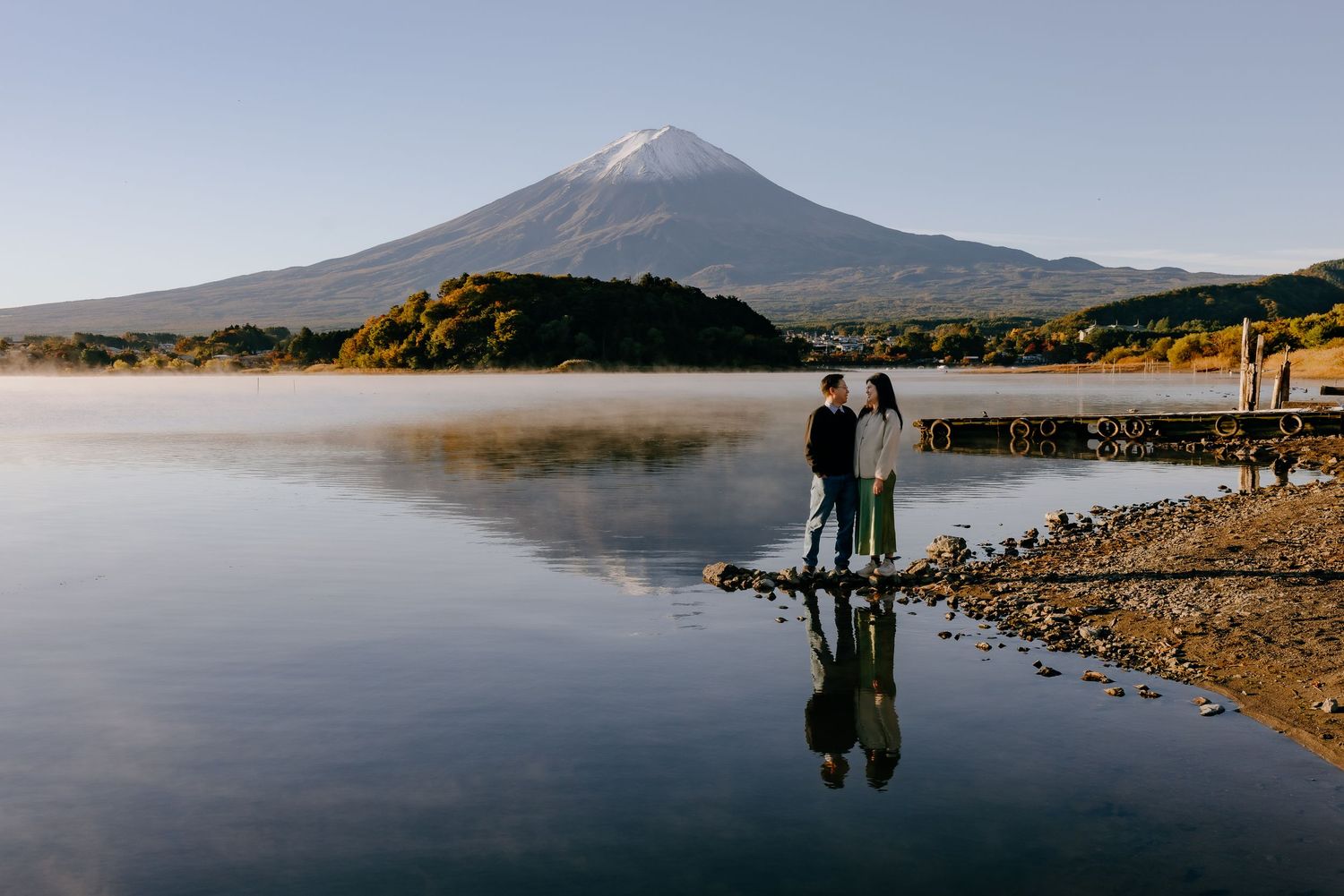 Silhouettes stand by a misty lake with a majestic mountain peak in the background at sunset.