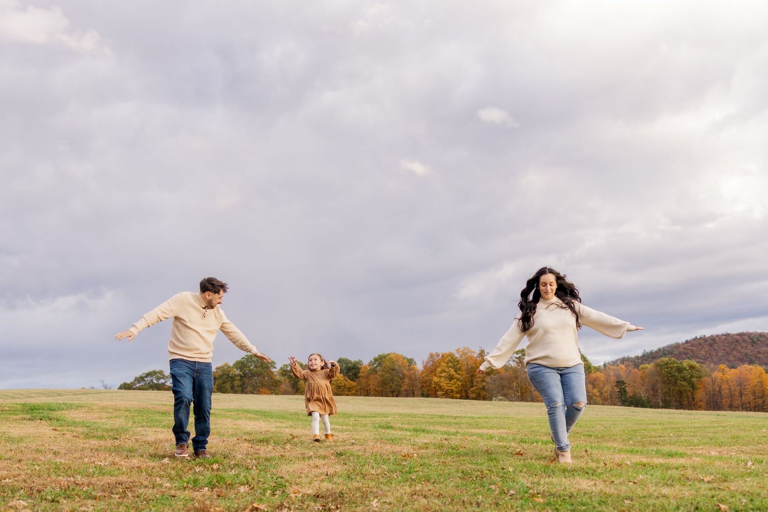 Two people run playfully through an autumn field with arms outstretched under a cloudy sky.